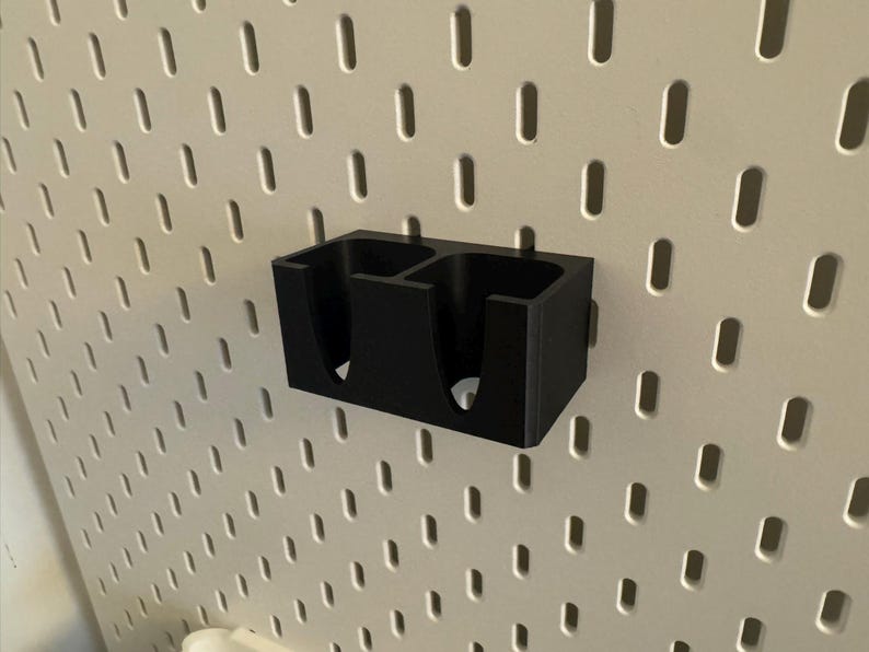 Puede incluir: Un organizador negro impreso en 3D con dos compartimentos, montado en una pared perforada blanca. El organizador es rectangular con aberturas redondeadas. La pared perforada tiene una cuadr&iacute;cula de agujeros espaciados.