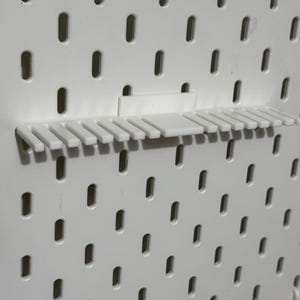 K&ouml;nnte beinhalten: Eine wei&szlig;e Lochwand mit einem wei&szlig;en Kunststoffregal, das daran befestigt ist. Das Regal hat 12 F&auml;cher zum Aufbewahren von Gegenst&auml;nden.