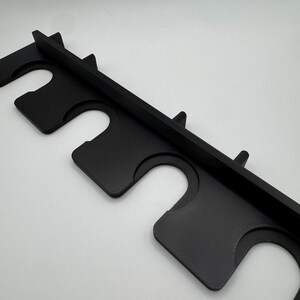 May include: A black, 3D-printed wall-mounted rack with multiple curved slots. The rack has a rectangular shape with a flat top and is designed to hold items. The material appears to be plastic.