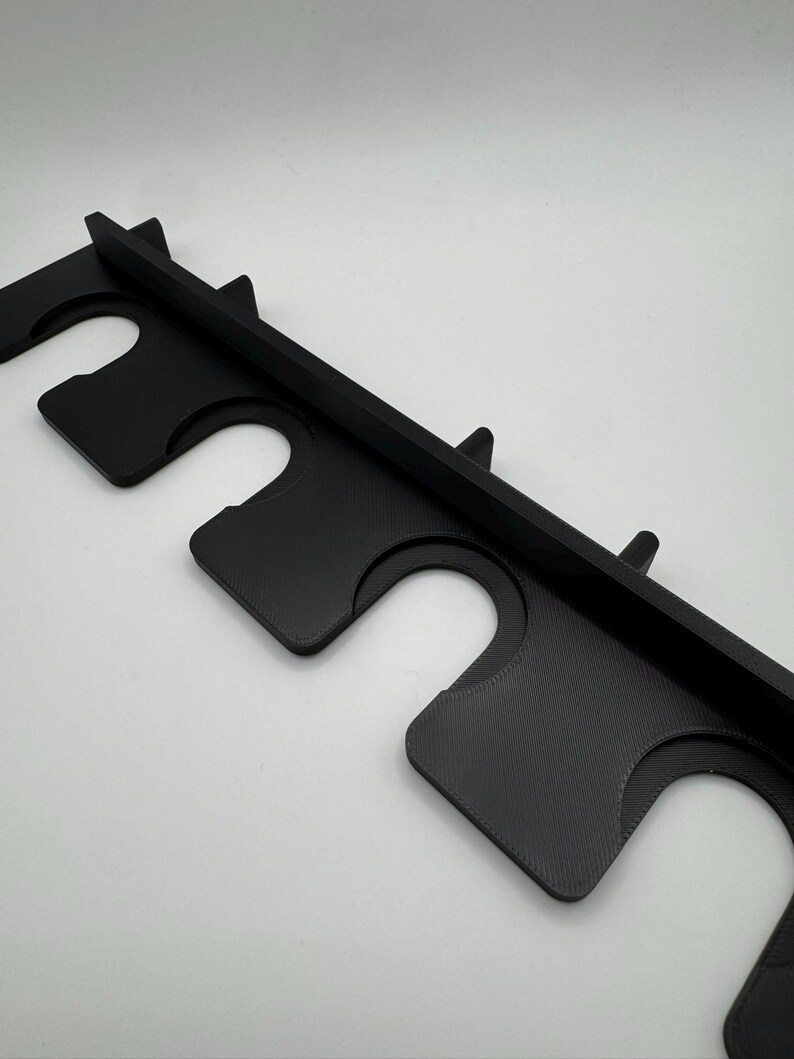 May include: A black, 3D-printed wall-mounted rack with multiple curved slots. The rack has a rectangular shape with a flat top and is designed to hold items. The material appears to be plastic.
