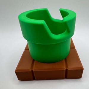 May include: A bright green pipe, with a distinctive curved opening, is placed on a brown, brick-like base. The design is reminiscent of a video game. The object is a three-dimensional representation of a classic game element.