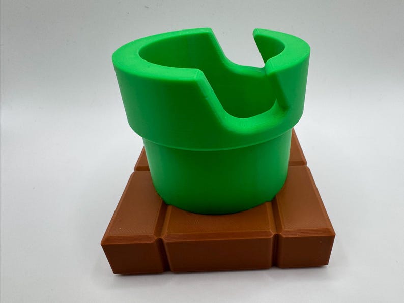 May include: A bright green pipe, with a distinctive curved opening, is placed on a brown, brick-like base. The design is reminiscent of a video game. The object is a three-dimensional representation of a classic game element.