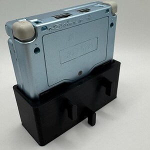 Pu&ograve; includere: Una console per videogiochi portatile Game Boy Advance SP di colore azzurro chiaro &egrave; montata su un supporto stampato in 3D nero. Il supporto ha due supporti laterali e una base.