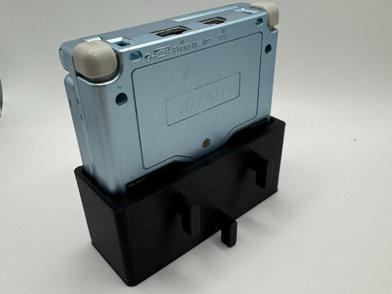 Pu&ograve; includere: Una console per videogiochi portatile Game Boy Advance SP di colore azzurro chiaro &egrave; montata su un supporto stampato in 3D nero. Il supporto ha due supporti laterali e una base.