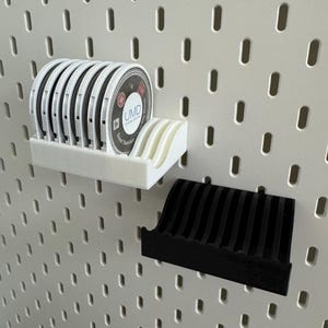 以下が含まれることがあります： 白い3DプリントのUMDディスクホルダーとUMDディスクの列。黒い3Dプリントのホルダーが下にあります。ホルダーは白いペグボードに取り付けられています。UMDディスクには白黒のラベルが付いています。