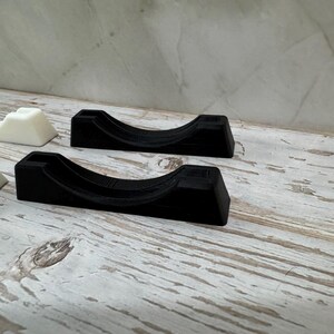 Puede incluir: Varios soportes negros y blancos impresos en 3D est&aacute;n dispuestos sobre una superficie de madera envejecida y blanqueada. Los soportes tienen un dise&ntilde;o curvo, con una base rectangular y una parte superior redondeada. Es probable que los soportes est&eacute;n dise&ntilde;ados para sujetar un objeto.