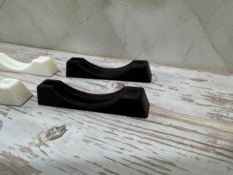 Puede incluir: Varios soportes negros y blancos impresos en 3D est&aacute;n dispuestos sobre una superficie de madera envejecida y blanqueada. Los soportes tienen un dise&ntilde;o curvo, con una base rectangular y una parte superior redondeada. Es probable que los soportes est&eacute;n dise&ntilde;ados para sujetar un objeto.