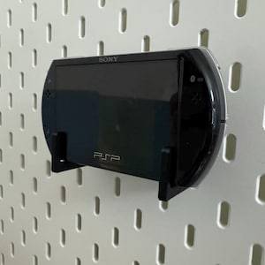 May include: Black Sony PSP handheld game console mounted on a white pegboard. The console is held in place by a black bracket. The PSP logo is visible, along with the Sony brand name.