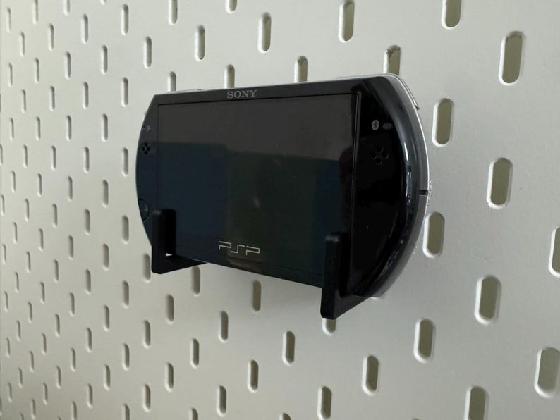 May include: Black Sony PSP handheld game console mounted on a white pegboard. The console is held in place by a black bracket. The PSP logo is visible, along with the Sony brand name.