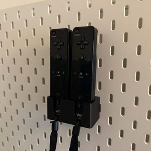 Puede incluir: Dos mandos Wii negros montados en una pared perforada blanca. Los mandos est&aacute;n en una base de carga negra. Los mandos tienen un mando direccional en forma de cruz y otros botones. Cuerdas negras cuelgan de la parte inferior de los mandos.