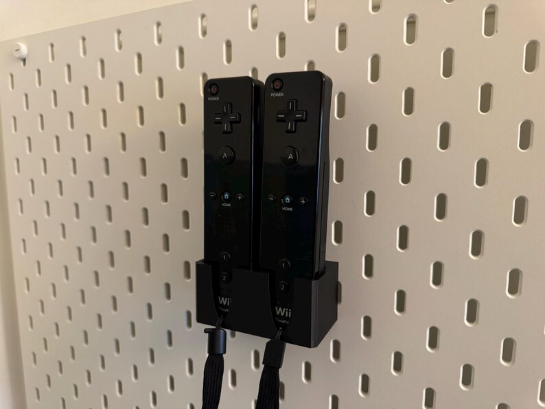 Puede incluir: Dos mandos Wii negros montados en una pared perforada blanca. Los mandos est&aacute;n en una base de carga negra. Los mandos tienen un mando direccional en forma de cruz y otros botones. Cuerdas negras cuelgan de la parte inferior de los mandos.