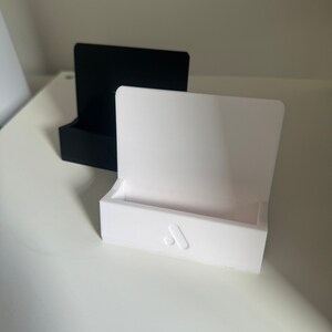 May include: Two minimalist phone stands, one black and one white, are displayed on a white surface. The white stand features a subtle logo on the front. The stands have a rectangular base and a curved back.