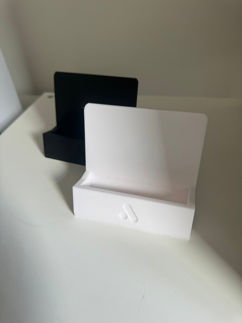 May include: Two minimalist phone stands, one black and one white, are displayed on a white surface. The white stand features a subtle logo on the front. The stands have a rectangular base and a curved back.