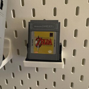 May include: A Nintendo Game Boy cartridge for "The Legend of Zelda: Link's Awakening" is displayed on a white pegboard. The cartridge is gray with a yellow and red label. The game is mounted on a black and white shelf.