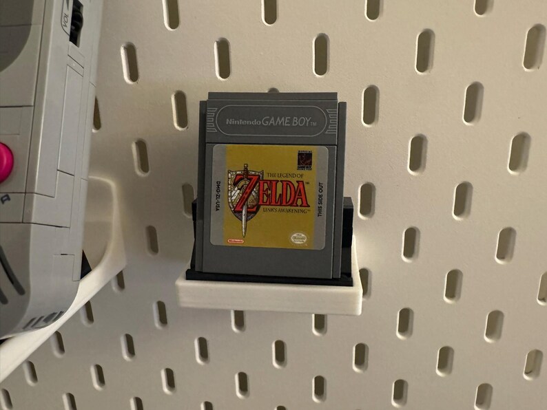 May include: A Nintendo Game Boy cartridge for "The Legend of Zelda: Link's Awakening" is displayed on a white pegboard. The cartridge is gray with a yellow and red label. The game is mounted on a black and white shelf.