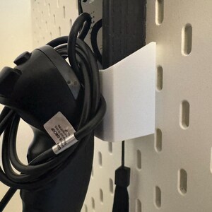May include: A black game controller with a coiled black cable is held by a white bracket attached to a pegboard. The pegboard is off-white with evenly spaced, vertical slots. A black tassel hangs below the bracket.