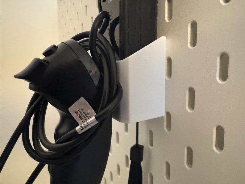 May include: A black game controller with a coiled black cable is held by a white bracket attached to a pegboard. The pegboard is off-white with evenly spaced, vertical slots. A black tassel hangs below the bracket.