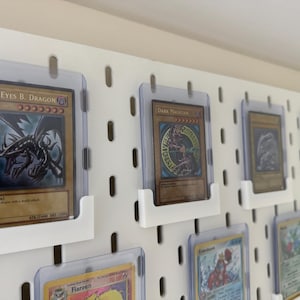 Pu&ograve; includere: Un pannello perforato bianco con diversi porta carte in plastica trasparente che mostrano varie carte collezionabili. Le carte presentano illustrazioni di creature e personaggi fantastici, tra cui "Red-Eyes B. Dragon" e "Dark Magician".