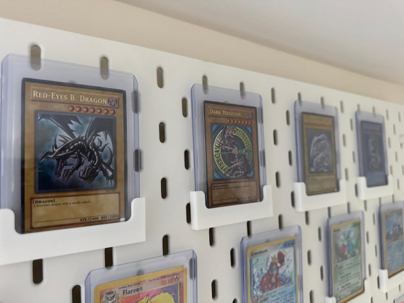 Pu&ograve; includere: Un pannello perforato bianco con diversi porta carte in plastica trasparente che mostrano varie carte collezionabili. Le carte presentano illustrazioni di creature e personaggi fantastici, tra cui "Red-Eyes B. Dragon" e "Dark Magician".