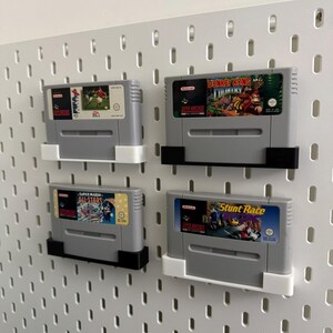 May include: Four grey Super Nintendo game cartridges displayed on a white pegboard. The cartridges are held in place by black and white holders. The game labels feature colourful graphics and titles such as "Super Mario All-Stars" and "Donkey Kong Country."