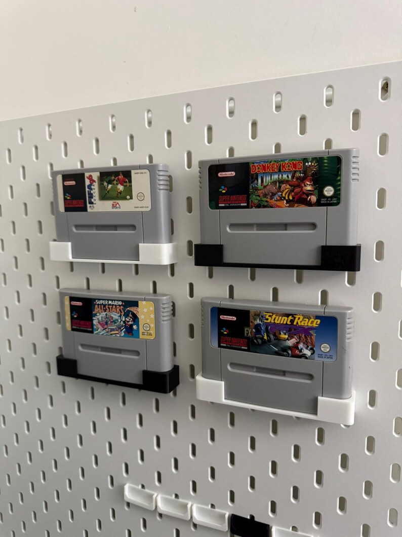 May include: Four grey Super Nintendo game cartridges displayed on a white pegboard. The cartridges are held in place by black and white holders. The game labels feature colourful graphics and titles such as "Super Mario All-Stars" and "Donkey Kong Country."