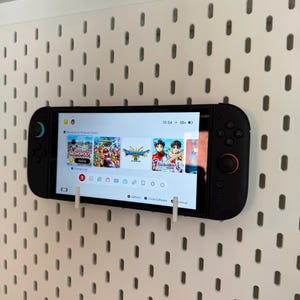 May include: A black Nintendo Switch console with attached controllers is mounted on a white pegboard. The screen displays game selection options. The time is 10:54.