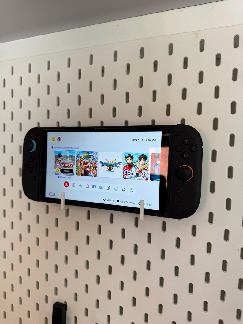 May include: A black Nintendo Switch console with attached controllers is mounted on a white pegboard. The screen displays game selection options. The time is 10:54.