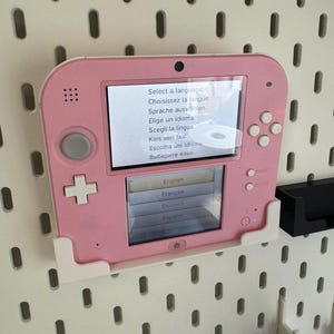 May include: A pink Nintendo 2DS handheld game console is mounted on a white pegboard. The screen displays a language selection menu with options in English, French, German, and Spanish. The console has a D-pad, buttons, and a small camera.