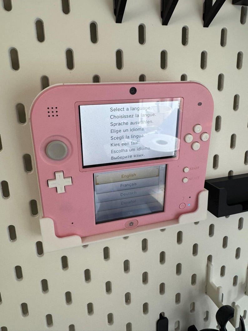 May include: A pink Nintendo 2DS handheld game console is mounted on a white pegboard. The screen displays a language selection menu with options in English, French, German, and Spanish. The console has a D-pad, buttons, and a small camera.