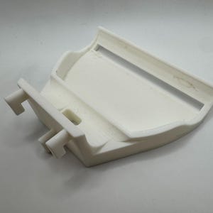 May include: A white, 3D-printed plastic object with a curved design. It features a rectangular opening and two protruding, rectangular tabs. The object appears to be a component or part of a larger assembly.