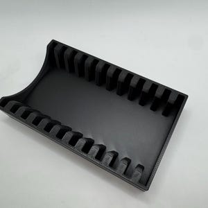 Op de afbeelding: Een zwarte rechthoekige organizer met meerdere verticale sleuven. De organizer heeft een gebogen rand aan &eacute;&eacute;n kant en is ontworpen om items vast te houden en weer te geven. Het product is gemaakt van een matzwart materiaal.