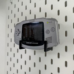 May include: A silver Game Boy Advance console mounted on a white pegboard. The handheld console is held by a black bracket. The screen is black, and the text "GAME BOY ADVANCE" is visible.