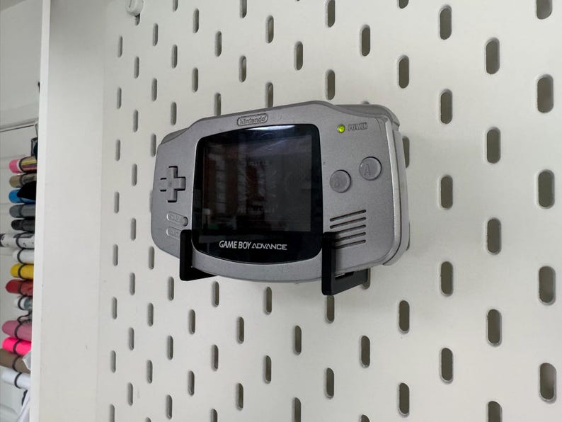 May include: A silver Game Boy Advance console mounted on a white pegboard. The handheld console is held by a black bracket. The screen is black, and the text "GAME BOY ADVANCE" is visible.