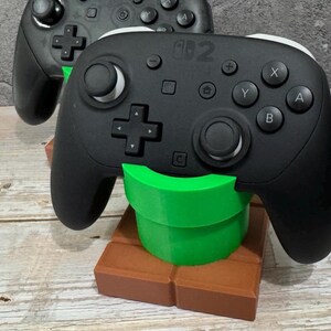 K&ouml;nnte beinhalten: Schwarzer Switch Pro Controller, der auf einem gr&uuml;n-braunen 3D-gedruckten St&auml;nder ruht. Der St&auml;nder ist so gestaltet, dass er einem gr&uuml;nen Rohr und einem braunen Ziegelstein aus einem beliebten Videospiel &auml;hnelt. Der Controller hat wei&szlig;e Akzente und verschiedene Tasten.