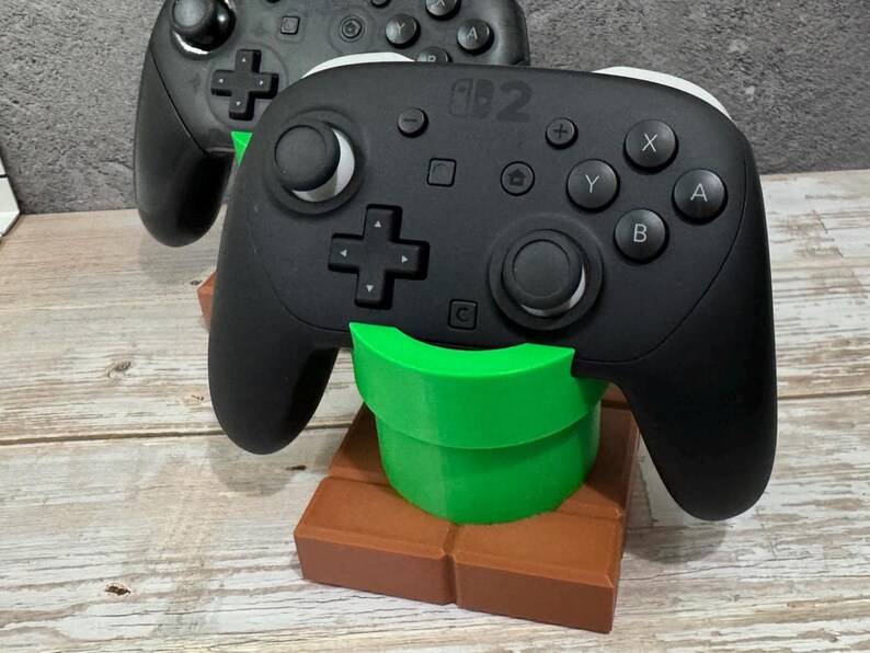K&ouml;nnte beinhalten: Schwarzer Switch Pro Controller, der auf einem gr&uuml;n-braunen 3D-gedruckten St&auml;nder ruht. Der St&auml;nder ist so gestaltet, dass er einem gr&uuml;nen Rohr und einem braunen Ziegelstein aus einem beliebten Videospiel &auml;hnelt. Der Controller hat wei&szlig;e Akzente und verschiedene Tasten.