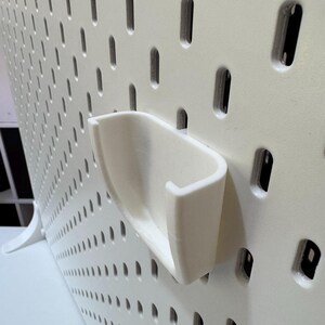 K&ouml;nnte beinhalten: Eine wei&szlig;e Lochwand mit einem befestigten wei&szlig;en Haken. Die Lochwand hat zahlreiche kleine, gleichm&auml;&szlig;ig verteilte L&ouml;cher. Der Haken ist so konzipiert, dass er in die L&ouml;cher passt und eine Aufbewahrungsl&ouml;sung bietet. Die Lochwand und der Haken bestehen aus einem soliden, langlebigen Material.
