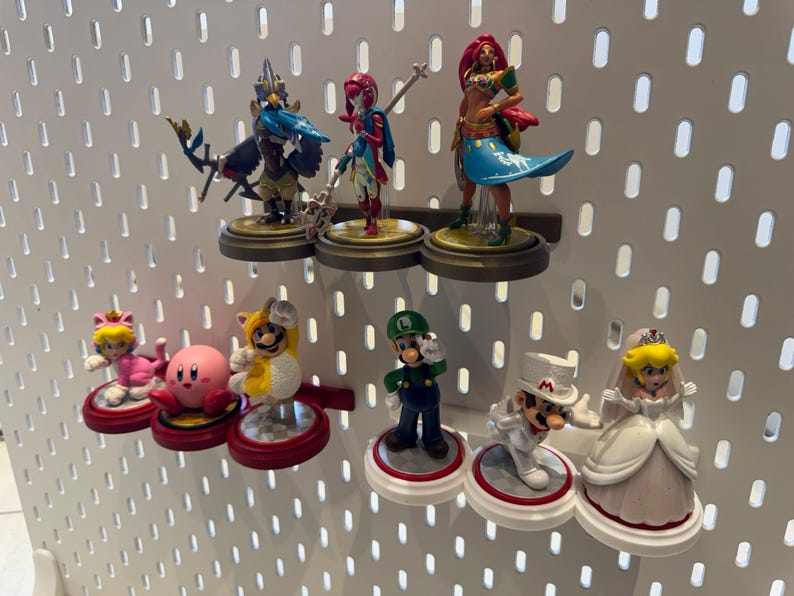 May include: A collection of colorful figurines displayed on a white pegboard. The figurines include characters from popular video games, such as Mario, Zelda, and Kirby. The figurines are displayed on small, round platforms.