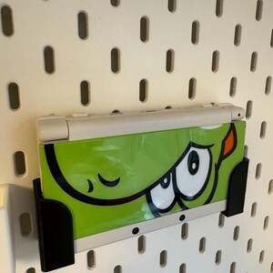 Puede incluir: Consola de juegos port&aacute;til Nintendo 3DS blanca y verde con un gr&aacute;fico de dibujos animados de un monstruo verde. La consola est&aacute; montada en una pared perforada blanca con un soporte negro.