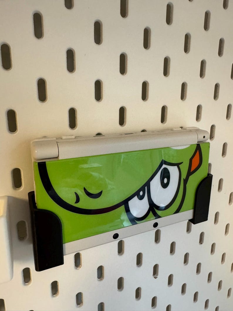 Puede incluir: Consola de juegos port&aacute;til Nintendo 3DS blanca y verde con un gr&aacute;fico de dibujos animados de un monstruo verde. La consola est&aacute; montada en una pared perforada blanca con un soporte negro.