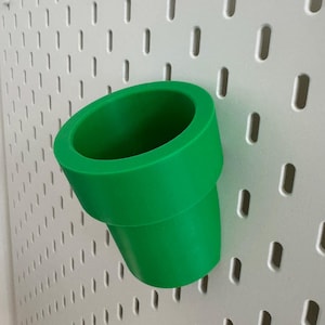 May include: A bright green, cylindrical pipe-shaped object, possibly a decorative item or a holder, is mounted on a white pegboard. The pipe has a wide opening and a slightly tapered base. The pegboard has a grid of evenly spaced, oblong holes.