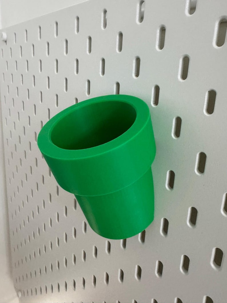 May include: A bright green, cylindrical pipe-shaped object, possibly a decorative item or a holder, is mounted on a white pegboard. The pipe has a wide opening and a slightly tapered base. The pegboard has a grid of evenly spaced, oblong holes.