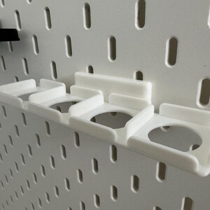 K&ouml;nnte beinhalten: Wei&szlig;e 3D-gedruckte Lochwand-Organizer. Der Haupt-Organizer hat drei kreisf&ouml;rmige &Ouml;ffnungen und ist an einer wei&szlig;en Lochwand befestigt. Ein kleinerer schwarzer Organizer ist ebenfalls an der Lochwand befestigt. Die Lochwand hat gleichm&auml;&szlig;ig verteilte ovale L&ouml;cher.