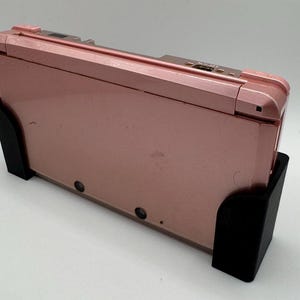 Pode incluir: Um console de jogos port&aacute;til Nintendo 3DS rosa em um suporte preto. O console est&aacute; fechado e o suporte &eacute; de pl&aacute;stico.