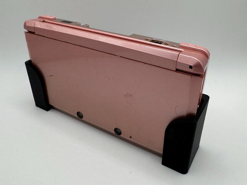 Pode incluir: Um console de jogos port&aacute;til Nintendo 3DS rosa em um suporte preto. O console est&aacute; fechado e o suporte &eacute; de pl&aacute;stico.