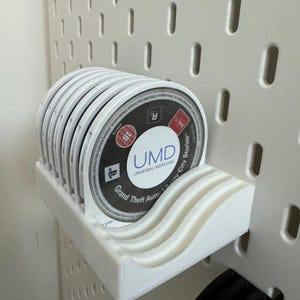 以下が含まれることがあります： 白い3DプリントのUMDディスクホルダーがペグボードに取り付けられ、いくつかのゲームディスクを保持しています。ディスクには「Grand Theft Auto Liberty City Stories」と「UMD UNIVERSAL MEDIA DISC」と記載されています。