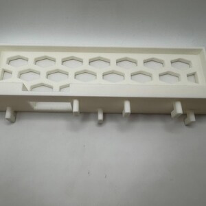 Puede incluir: Un objeto rectangular de pl&aacute;stico blanco con un patr&oacute;n de panal y patas de soporte. El objeto tiene un dise&ntilde;o tipo bandeja con una serie de recortes hexagonales. Las patas est&aacute;n espaciadas a lo largo del borde inferior.