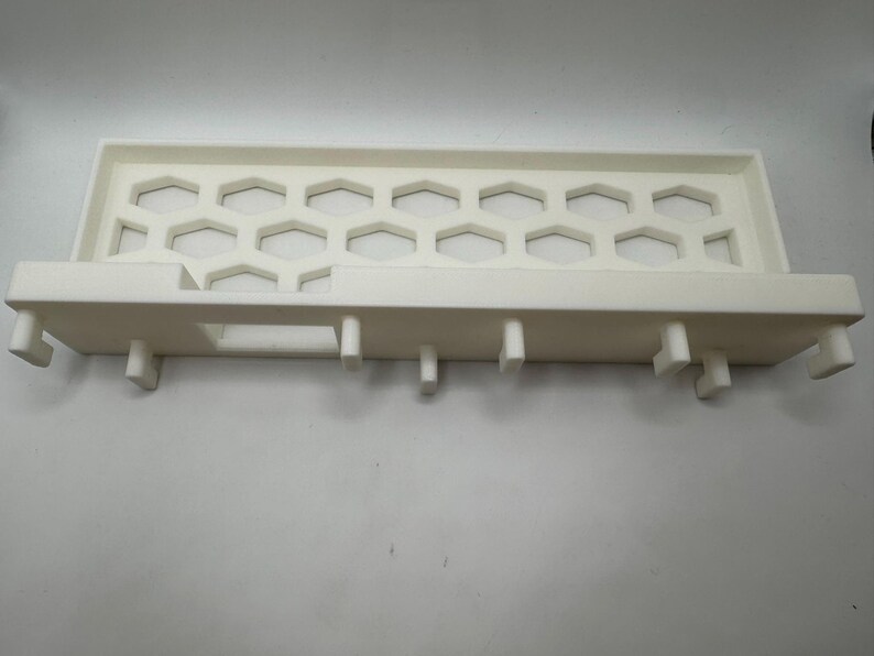 Puede incluir: Un objeto rectangular de pl&aacute;stico blanco con un patr&oacute;n de panal y patas de soporte. El objeto tiene un dise&ntilde;o tipo bandeja con una serie de recortes hexagonales. Las patas est&aacute;n espaciadas a lo largo del borde inferior.