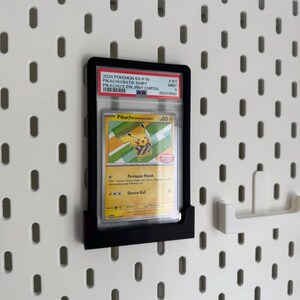 May include: A black display frame holding a Pokemon trading card featuring Pikachu. The card is encased in a protective sleeve and is mounted on a white pegboard wall. The card's details include the text "2024 Pokemon SV-P in Pikachu Batik Shirt".