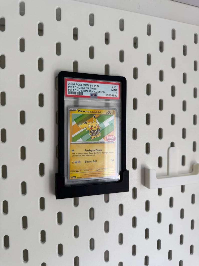 May include: A black display frame holding a Pokemon trading card featuring Pikachu. The card is encased in a protective sleeve and is mounted on a white pegboard wall. The card's details include the text "2024 Pokemon SV-P in Pikachu Batik Shirt".