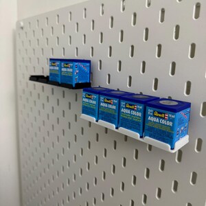 K&ouml;nnte beinhalten: Eine wei&szlig;e Lochwand mit mehreren kleinen blauen Schachteln mit Revell Aqua Color Acrylfarbe. Die Schachteln werden auf schwarzen und wei&szlig;en Regalen ausgestellt. Jede Schachtel enth&auml;lt 18 ml Farbe.